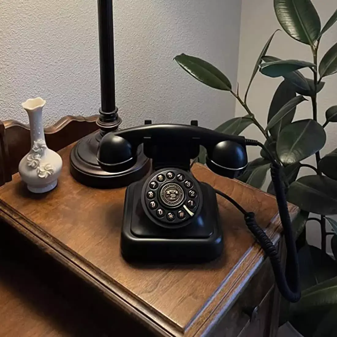 Telefon negru cu disc, pe o comodă de lemn, lângă o lampă și o vază albă